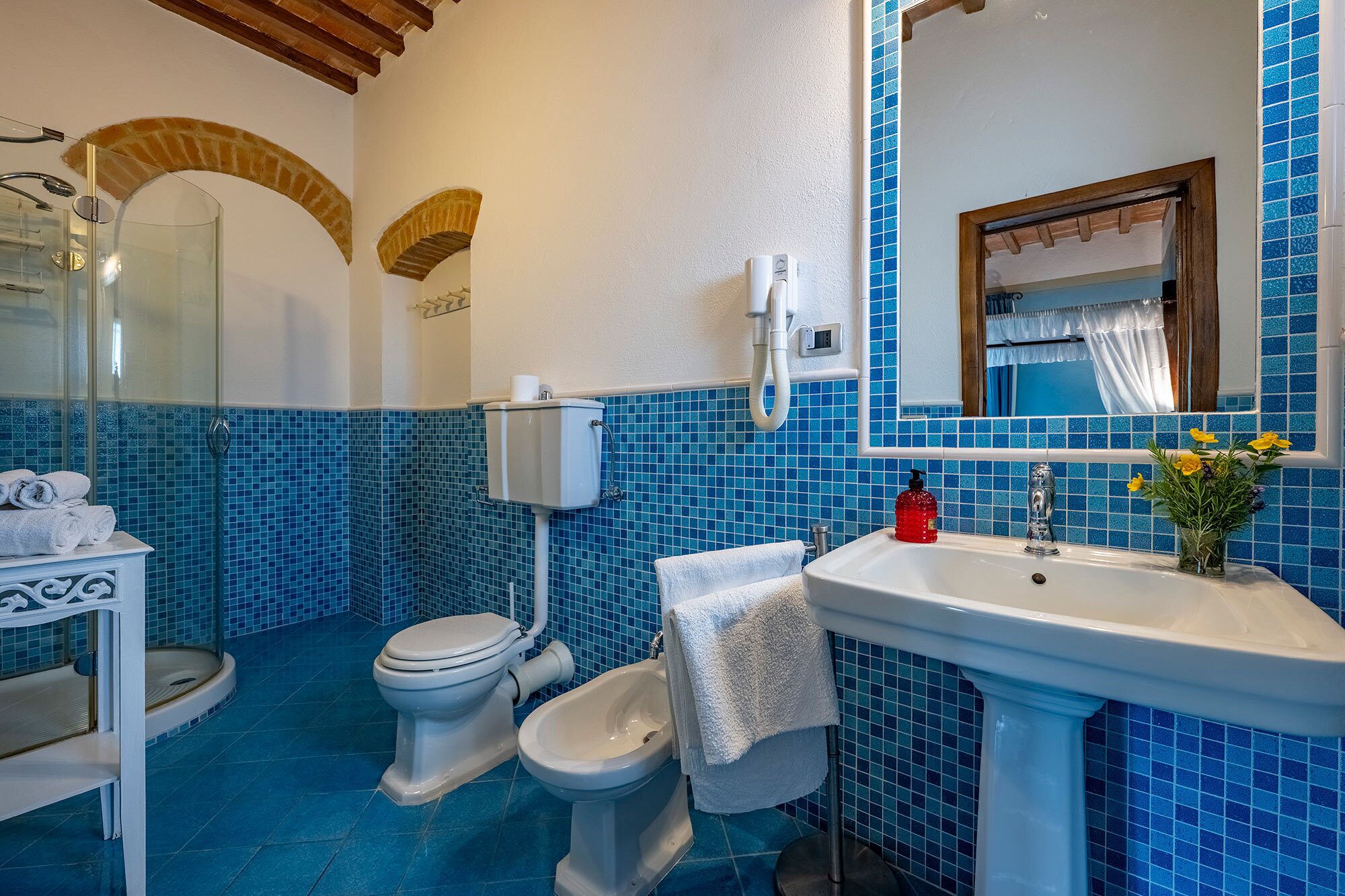 A bathroom with a toilet, bidet, and sink, featuring blue mosaic tiles and a wooden ceiling.