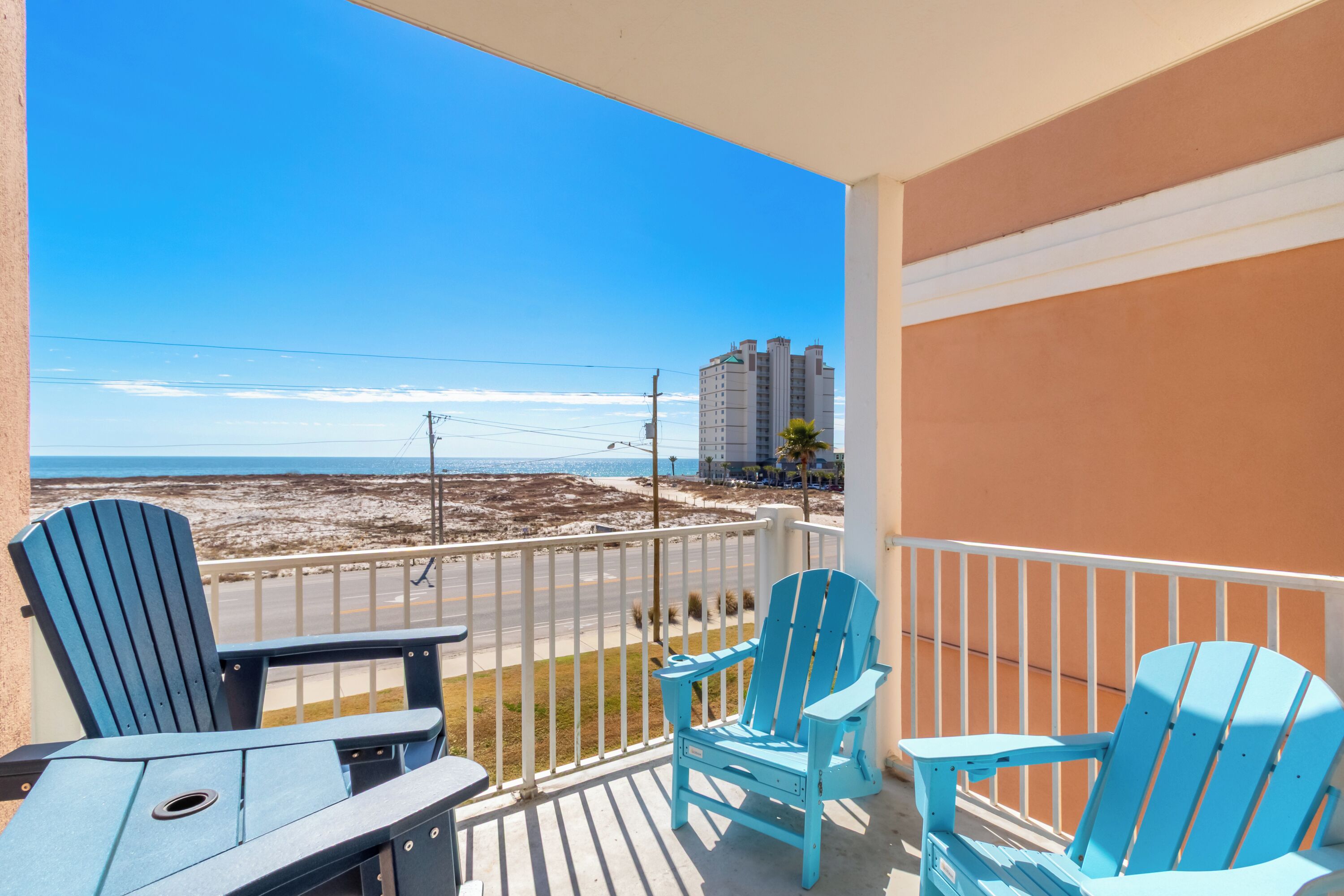A balcony with two blue Adirondack chairs and a small table, overlooking a beach with a clear sky.