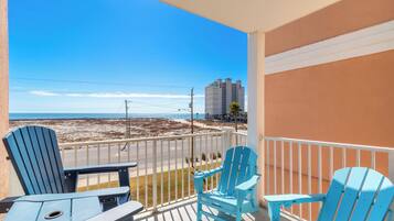 A balcony with two blue Adirondack chairs and a small table, overlooking a beach with a clear sky.