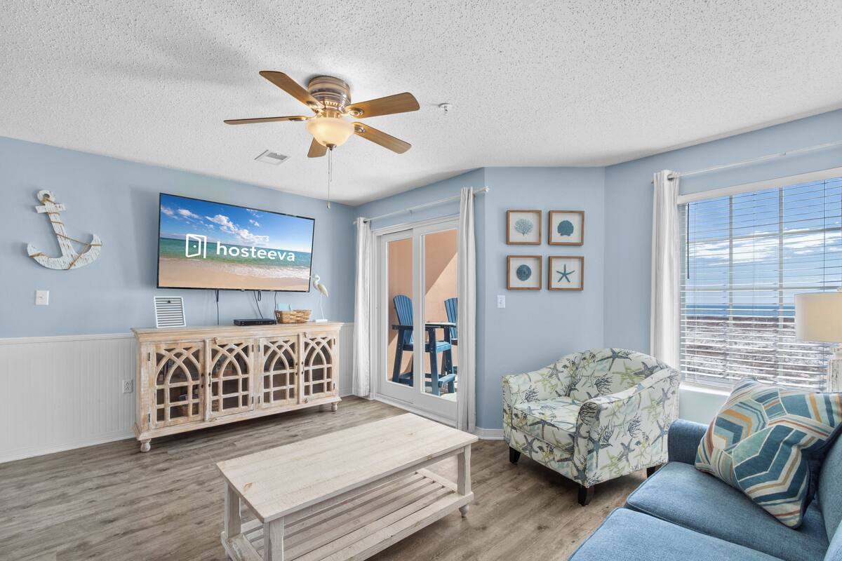 A living room with a ceiling fan, a television displaying 'hosteeva', a wooden cabinet, a patterned armchair, a sofa, and a view through a window.