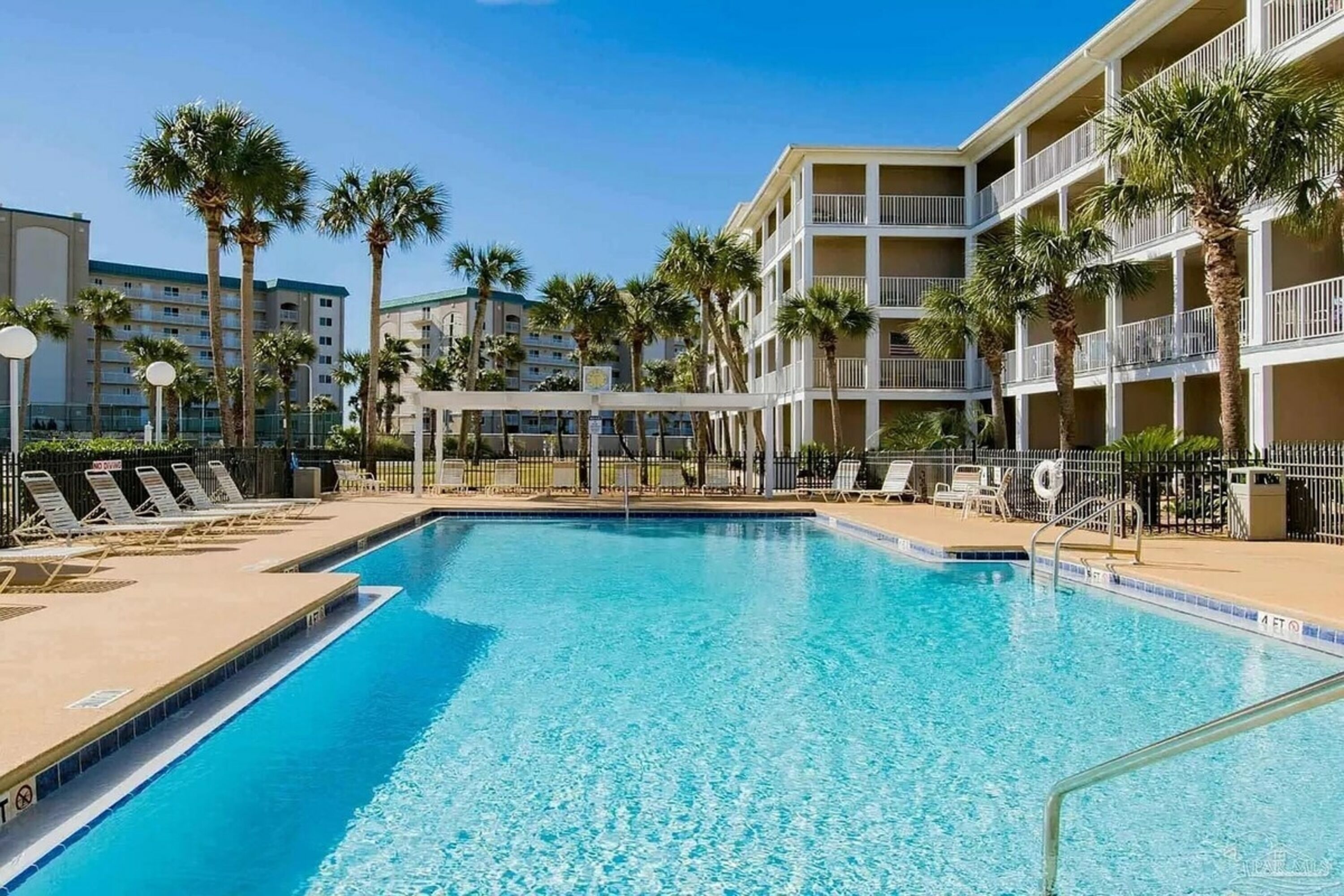 Ein Swimmingpool mit Liegestühlen und Palmen, umgeben von mehrstöckigen Gebäuden.