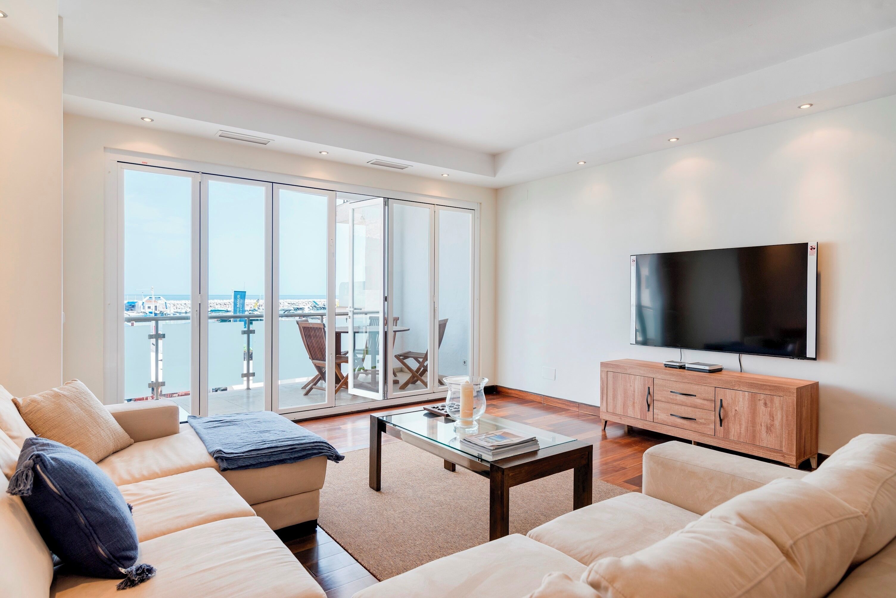 Een moderne woonkamer met een grote televisie, comfortabele banken, een glazen salontafel en uitzicht op het water en de skyline door grote schuifdeuren.