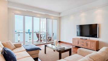 Een moderne woonkamer met een grote televisie, comfortabele banken, een glazen salontafel en uitzicht op het water en de skyline door grote schuifdeuren.