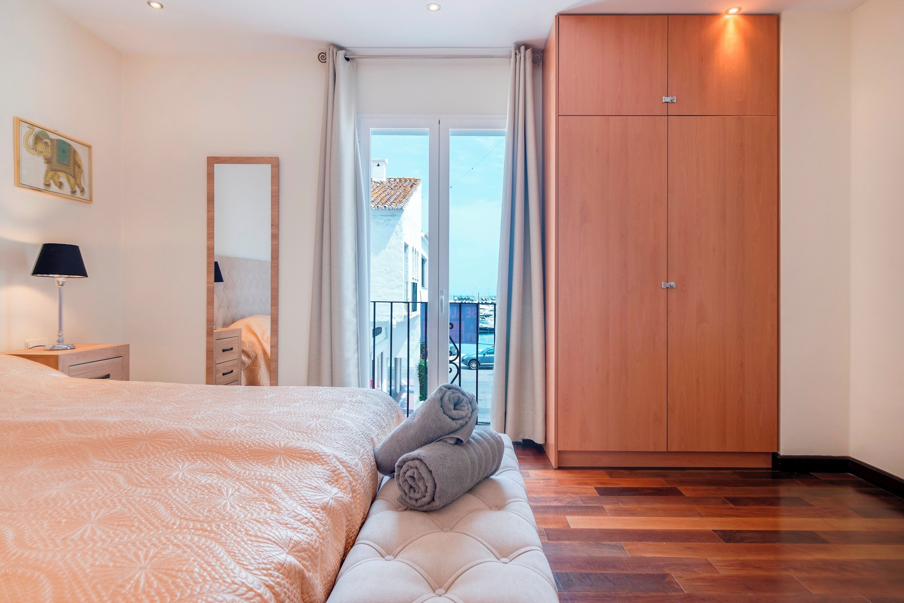 A bedroom with a large bed, a wooden wardrobe, a balcony with a view, and a painting on the wall.