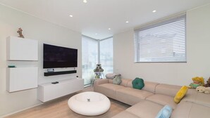 A modern living room with a flat-screen TV mounted on a white cabinet, a beige sofa with cushions, a round white ottoman, and a window with blinds. - Modern Smart Home with Garden, close to Richmond - Pass the Keys (London)