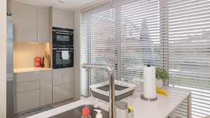 A modern kitchen with a built-in oven, a stainless steel sink, and a window with horizontal blinds. - Modern Smart Home with Garden, close to Richmond - Pass the Keys (London)