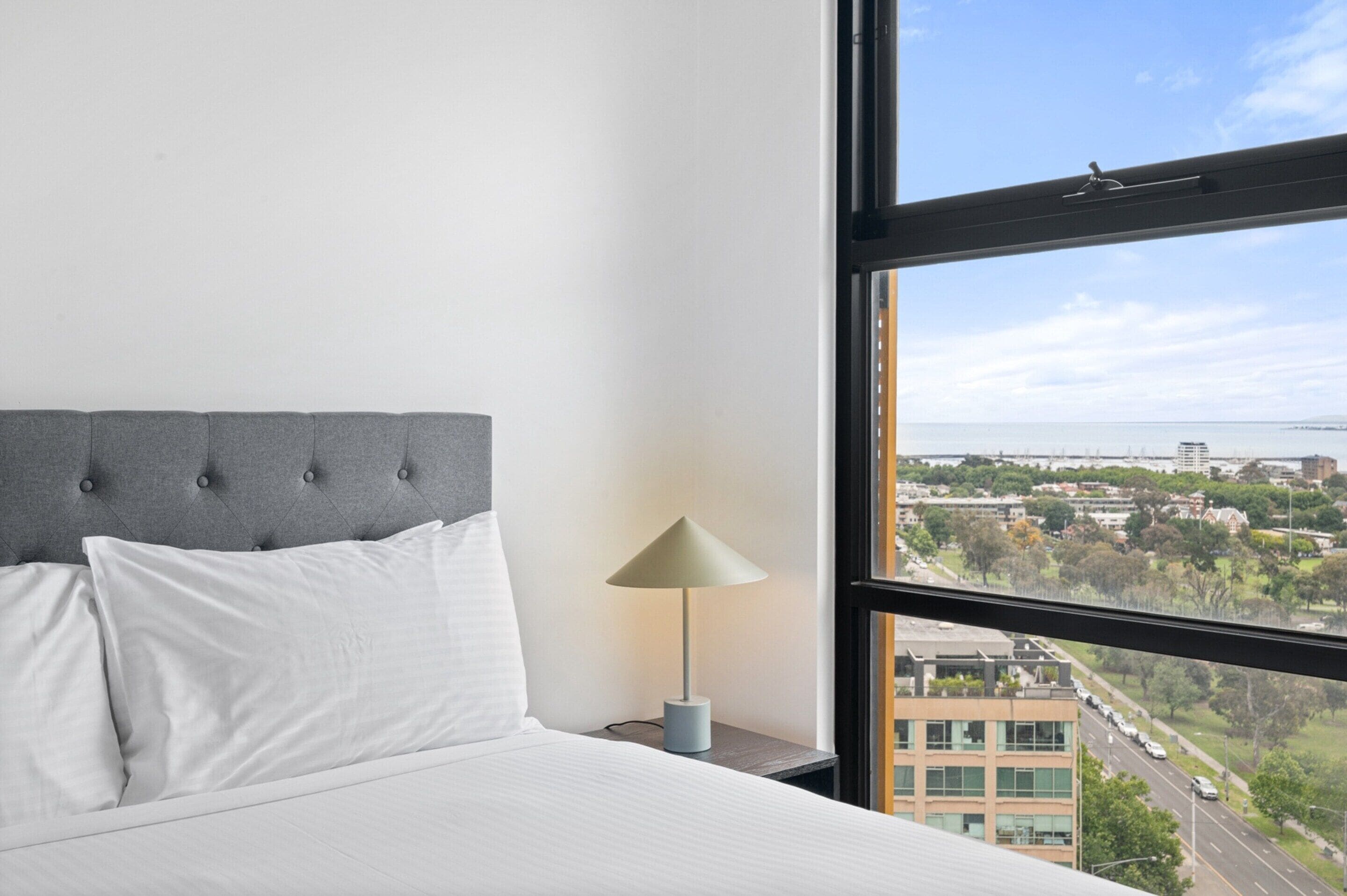 Ein Hotelzimmer mit großem Fenster, einem Bett mit weißer Bettwäsche, einem grau gepolsterten Kopfteil und einem Nachttisch mit Lampe.