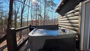 Un jacuzzi en una terraza de madera con vistas a un bosque y una cabaña de troncos al fondo.