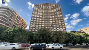 Un grand immeuble d’habitation doté de nombreux balcons, entouré de voitures garées et d’arbres.