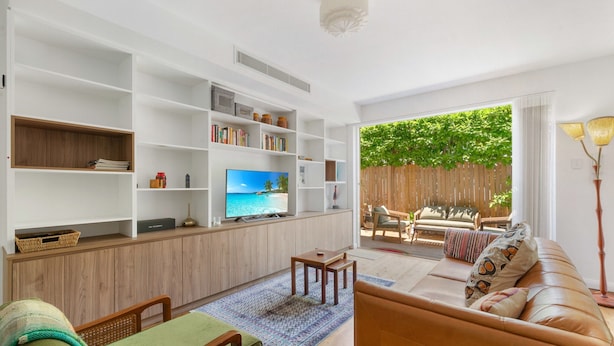 A living room with a TV, bookshelves, a sofa, and a view of a wooden fence and greenery. - Spacious Stay Near Shops & Cafes in Marrickville (Sydney)