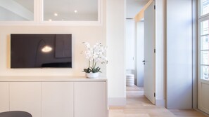 A modern interior with a flat-screen TV mounted on a wall, a potted orchid plant, and a hallway leading to a door. - Catarina 392 | Urban 1BR by LovelyStay (Porto)