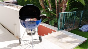 Ein tragbarer Grill mit geöffnetem Deckel, platziert auf einer Terrasse mit grünem Zaun und Pflanzen im Hintergrund.