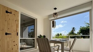 En rymlig balkong med ett matbord och stolar, stora fönster med utsikt över himlen och grönskan, och en skjutdörr i glas som leder in till ett inomhusområde.