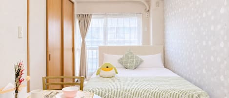 Un dormitorio con cama, una mesita de noche con un cuenco y una taza, una ventana con cortinas y un equipo de aire acondicionado.