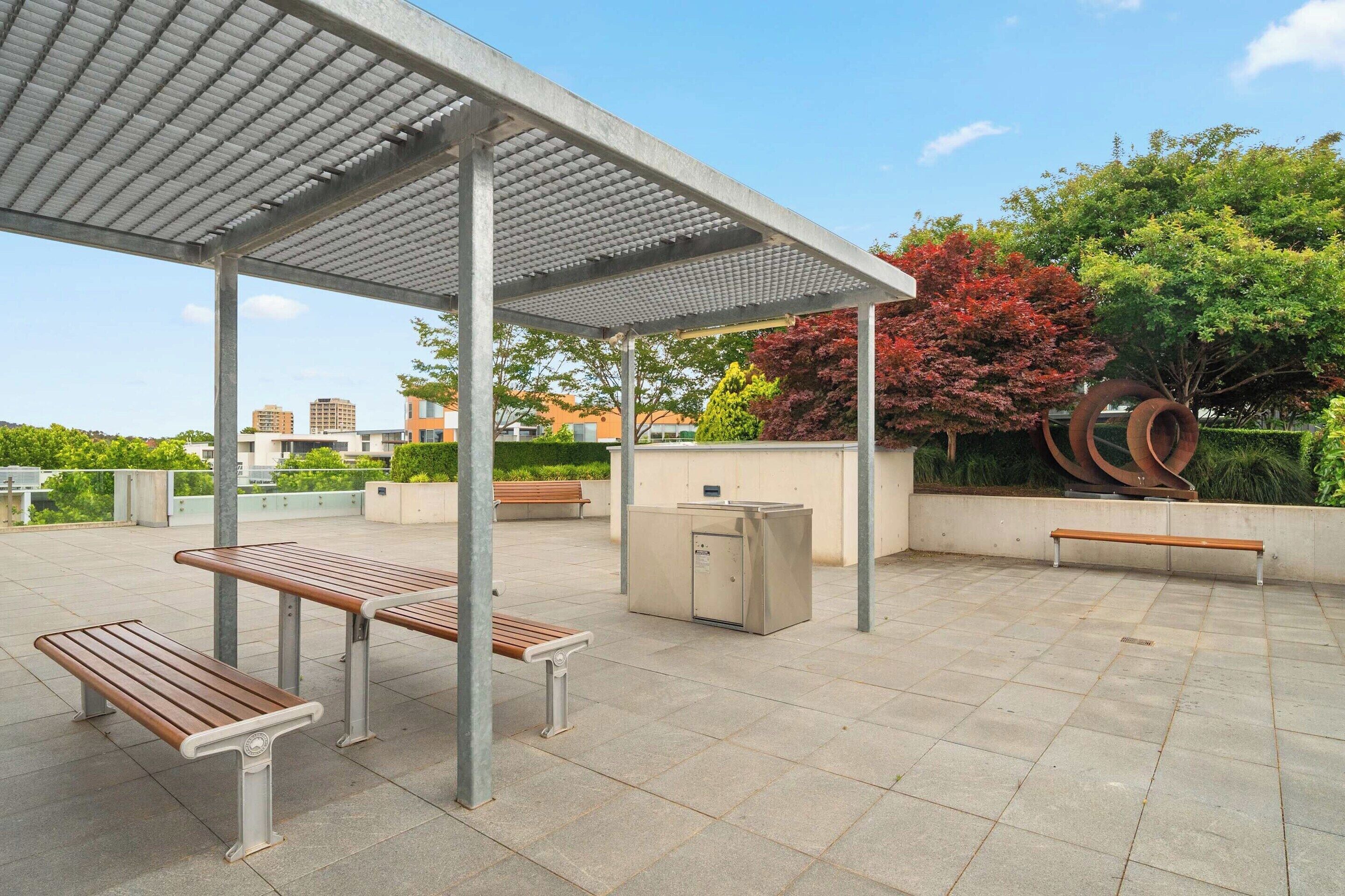 An outdoor seating area with benches and a covered shelter, surrounded by greenery and buildings.
