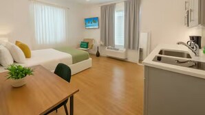 A modern hotel room with a bed, a small dining table with a plant, a kitchenette, and a window with curtains. - Miami Beach Quaint Studio  (Miami Beach)