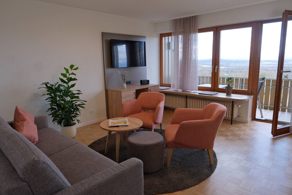 A modern living room with a sofa, two armchairs, a coffee table, a TV, a desk, and a large window with a view.
