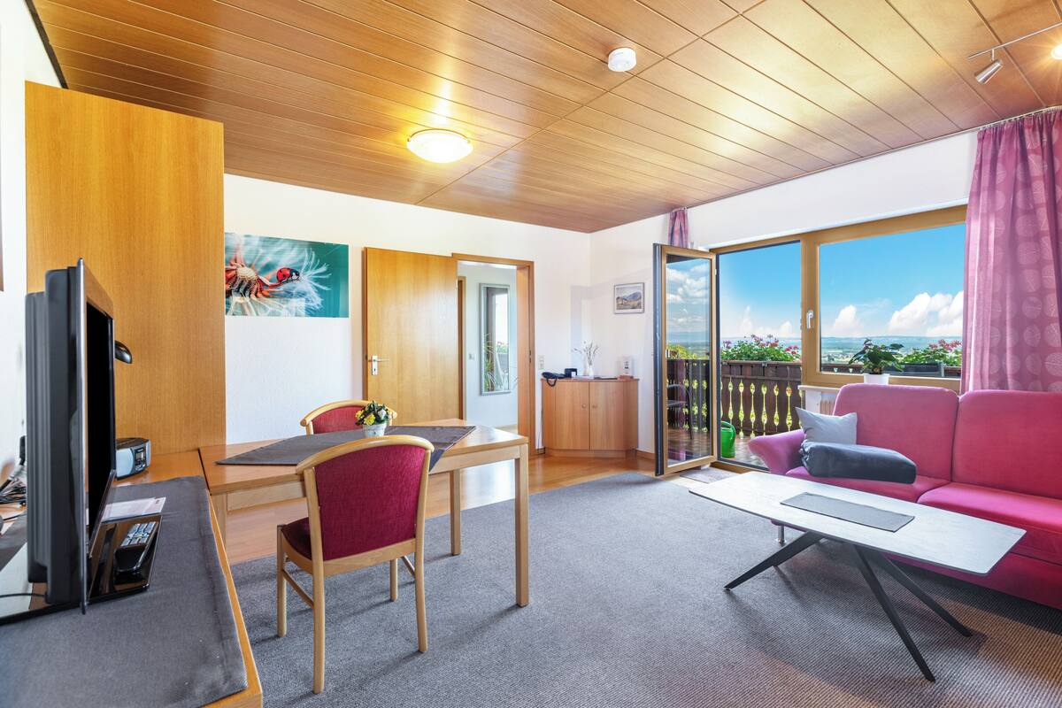 A modern room with a wooden ceiling, a dining area with a table and chairs, a pink sofa, and a large window offering a view.
