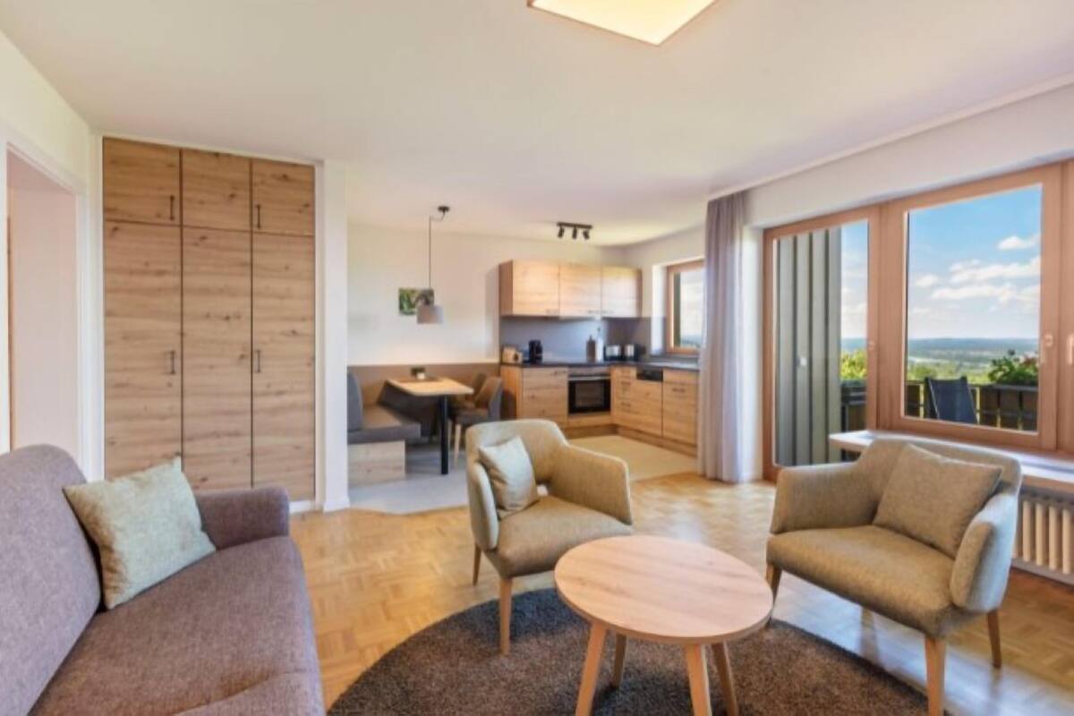 A modern living room with a sofa, two armchairs, a round coffee table, and a view of the kitchen and balcony.