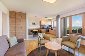 En moderne stue med sofa, to lænestole, et rundt sofabord og udsigt til køkken og balkon.