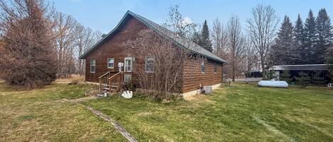 A rustic wooden cabin with a wooden staircase leading to a porch, surrounded by a grassy yard and trees.