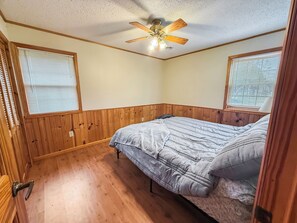 Un dormitorio con paneles de madera, una cama con colcha gris, un ventilador de techo y dos ventanas con persianas.