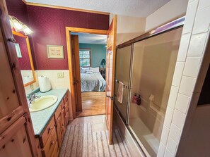 Un baño con tocador de madera, ducha y vista hacia un dormitorio con cama y ventana.