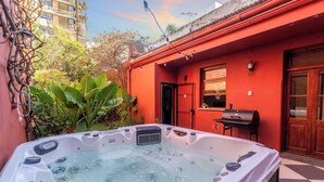 A hot tub in a courtyard with a red building, a grill, and lush greenery. - RARE Designer Loft w/ Garden, Jacuzzi & Rooftop! (DAI)
