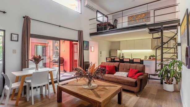 A modern living room with a dining area, a staircase leading to a loft, and a kitchen in the background. - RARE Designer Loft w/ Garden, Jacuzzi & Rooftop! (DAI)