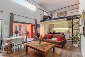 Una sala de estar moderna con comedor, una escalera que lleva a un altillo y una cocina al fondo.