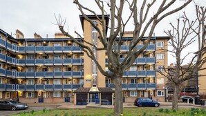 A multi-story residential building with balconies, a central entrance, and a small garden area with flowers. - 1 Bedroom flat in Trendy East London (London)