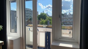 A view through a window with a white frame, revealing a balcony with a blue railing, a white pot, and a building in the background. - 1 Bedroom flat in Trendy East London (London)