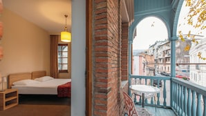 A bedroom with a bed, bedside table, and a window with curtains. It also shows a balcony with a table and chairs, overlooking a street with buildings and cars. - Tekla Palace Block B by Urban Hotels  (Tbilisi)