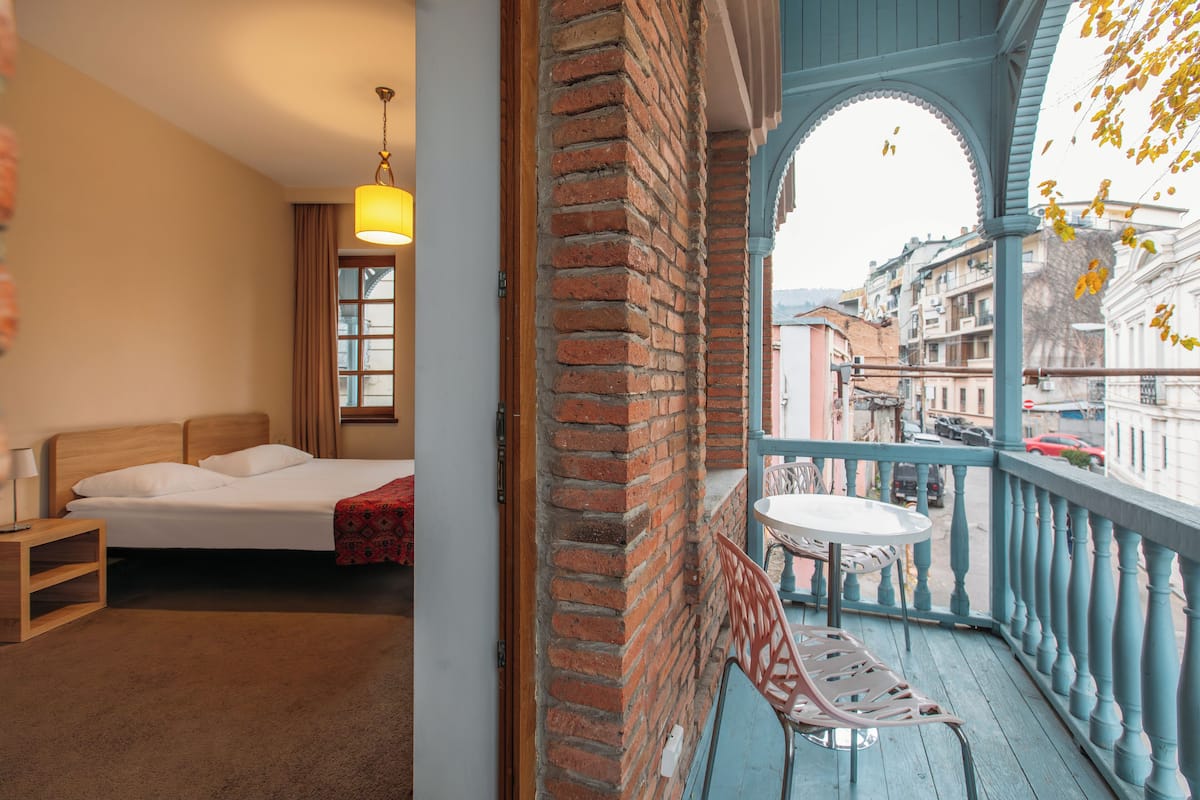A bedroom with a bed, bedside table, and a window with curtains. It also shows a balcony with a table and chairs, overlooking a street with buildings and cars.