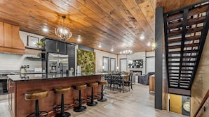 A modern kitchen with wooden ceiling beams, a central island with bar stools, stainless steel appliances, and a decorative wall with greenery. - Evergreen Escape - Luxury Rooftop - Indoor Pool - Home Theater - Game Rooms (Sevierville)
