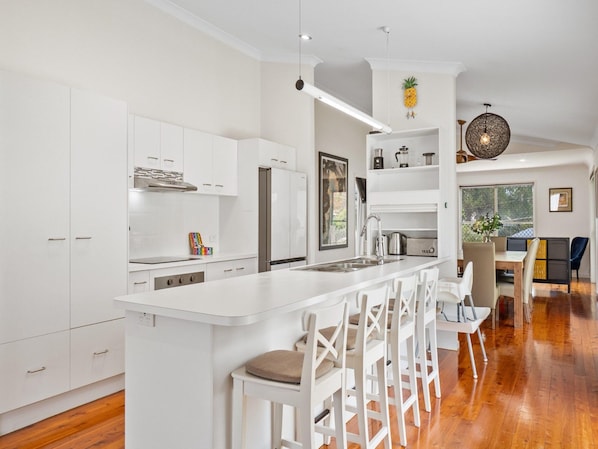 A modern kitchen with white cabinetry, a central island with bar stools, and a dining area with a view. - Bright 3-bed Home in Leafy Annerley (Brisbane)