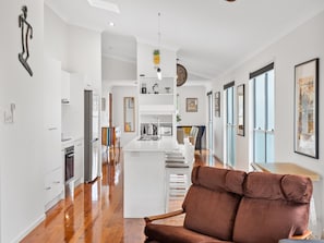A modern kitchen with white cabinetry, a central island, and a dining area with a brown upholstered chair. - Bright 3-bed Home in Leafy Annerley (Brisbane)