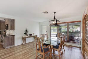 Una cocina amplia con armarios de madera, un comedor con mesa de vidrio y sillas de madera, y un gran ventanal con persianas.