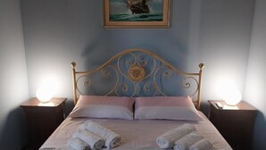 Un lit bien fait, avec des oreillers et des serviettes enroulées, une tête de lit décorative et un tableau représentant un navire accroché au mur.
