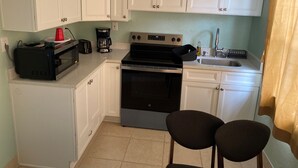 Een kleine keuken met witte kastjes, een fornuis, een spoelbak en twee stoelen.