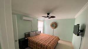 Een slaapkamer met een bed, een plafondventilator, een airconditioningapparaat, een tv aan de muur en een raam met gordijnen.