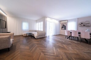 A modern living room with a herringbone wood floor, a sofa, a dining table with chairs, and a large window with sheer curtains.