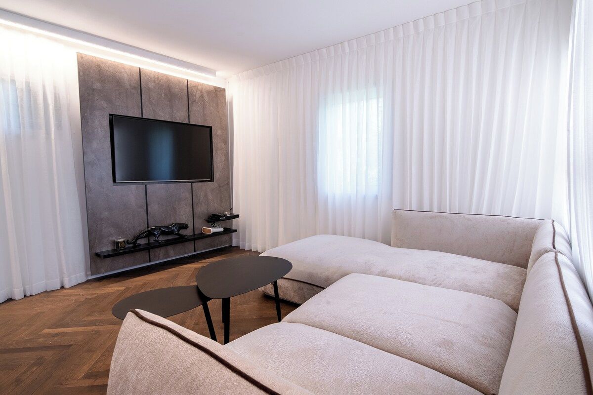 A modern living room with a flat-screen TV mounted on a wall, a sofa, a small table, and a herringbone-patterned wooden floor.