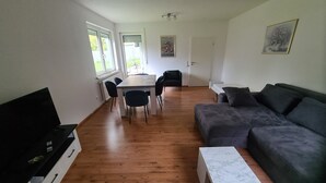 Een moderne woonkamer met een houten vloer, een grijze bank, een witte salontafel, een eettafel met stoelen en een zwart tv-meubel.