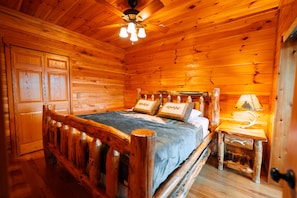 Un dormitorio de cabaña de madera con cama, mesita de noche, lámpara y ventilador de techo.