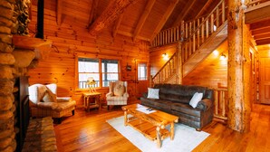 A log cabin interior with a wooden staircase, a leather sofa, plaid chairs, and a stone fireplace.
