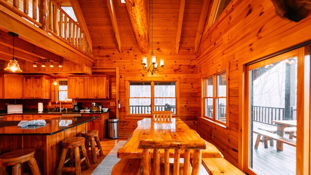A wooden cabin interior with a dining area, kitchen, and large windows.
