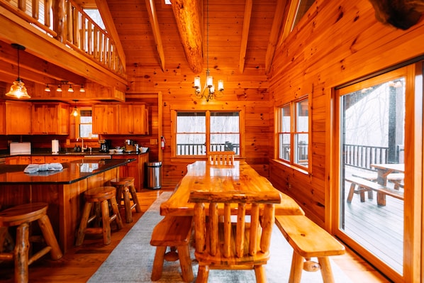 Interior de una cabaña de madera con comedor, cocina y ventanas amplias.