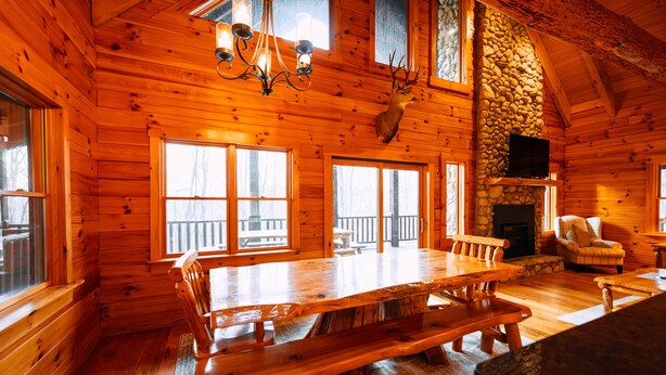 A log cabin interior with a large wooden dining table, benches, a stone fireplace, and a mounted deer head.