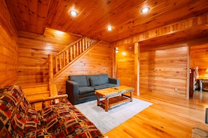 Interior d'una cabana de fusta amb escala, un sofà, una taula de centre i una llar de foc.